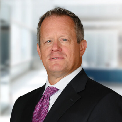 A middle-aged man in a suit and purple tie stands in front of a blurred law offices background, looking at the camera with a neutral expression.