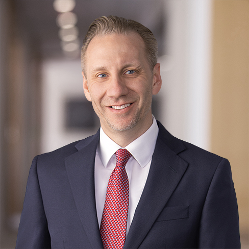 A man in a navy suit, white shirt, and red patterned tie is smiling, standing indoors with a blurred hallway in the background—capturing the professional image often seen at law offices of Chicago lawyers.