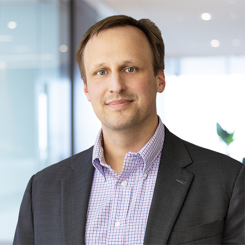 A man wearing a dark suit jacket and a checked dress shirt stands in a modern corporate law office, looking at the camera with a neutral expression.