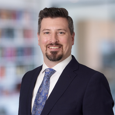 A man in a navy suit, white shirt, and patterned blue tie, smiling, stands in front of a blurred corporate law office background with shelves and books.