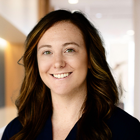 A woman with long brown hair and a dark top is smiling, standing in a brightly lit law offices setting, with a blurred background typical of Chicago lawyers' workspaces.