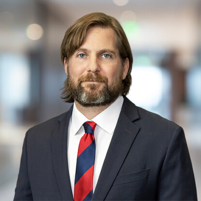 A man with shoulder-length brown hair and a beard, wearing a dark suit, white shirt, and red-and-blue striped tie, stands in the blurred backdrop of a bustling corporate law office.