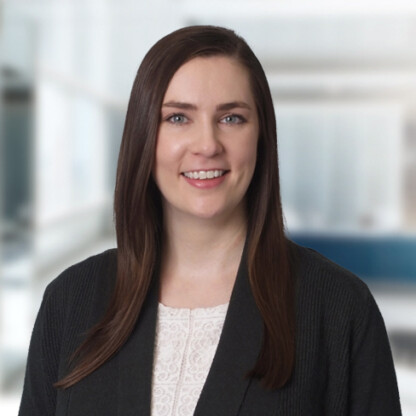 A woman with straight brown hair, wearing a white top and dark blazer, smiles at the camera in a bright, blurred corporate law office setting.