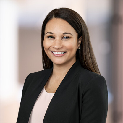 A woman with long brown hair, wearing a black blazer and light pink top, smiles at the camera against a blurred office background, reflecting the professionalism of Chicago lawyers in leading law offices.
