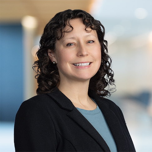 A woman with curly brown hair, wearing a black blazer and blue top, smiles at the camera against a blurred corporate law office background.