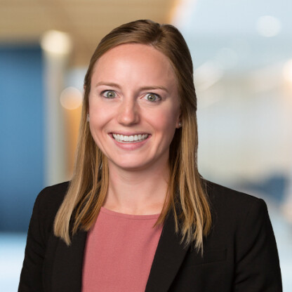 Woman with straight light brown hair wearing a black blazer and pink top, smiling at the camera in a blurred law offices setting, reflecting the professionalism of Chicago lawyers.