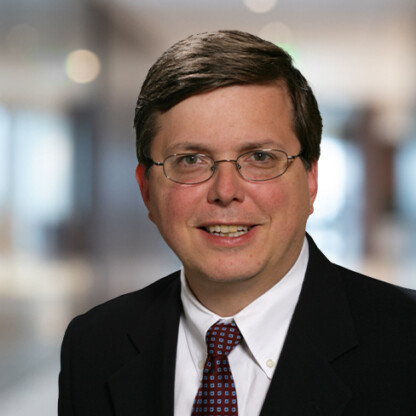 A man with short brown hair, glasses, and a dark suit smiles at the camera in a blurred indoor setting—reflecting the approachable professionalism of top lawyers in Chicago.