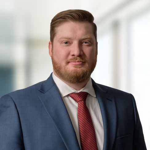 A man with short light brown hair and a beard wears a blue suit, white shirt, and red striped tie, standing in front of a blurred background at a corporate law office.