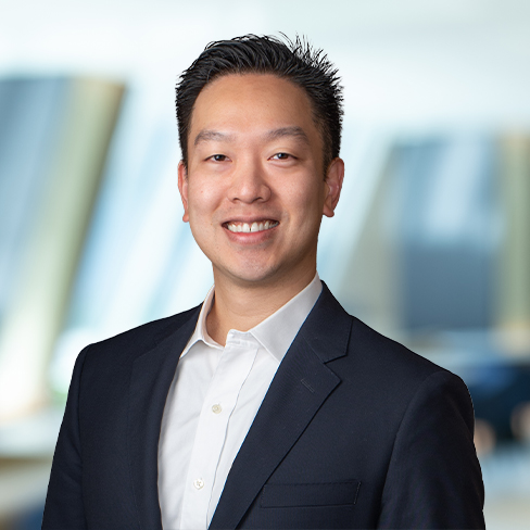 A man in a dark blazer and white shirt smiles at the camera, standing in front of a blurred indoor background with natural light, embodying the professionalism often found in law offices specializing in intellectual property law.