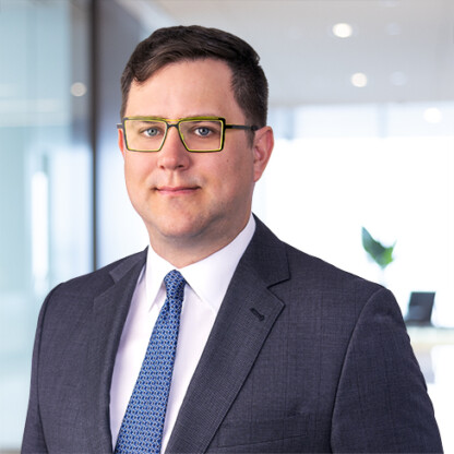 A man wearing glasses, a dark suit, white shirt, and blue patterned tie stands in a modern corporate law office with glass walls and bright lighting.