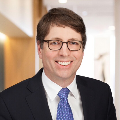 A man wearing glasses, a dark suit, white shirt, and patterned blue tie, smiling in a brightly lit corporate law office hallway.