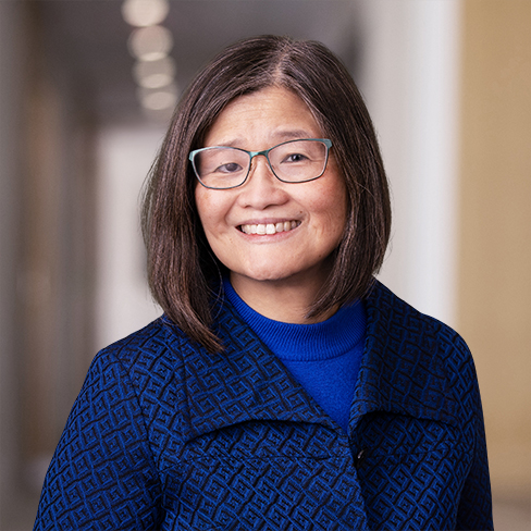A woman with shoulder-length brown hair and glasses smiles while wearing a blue patterned jacket in the hallway of a corporate law office.