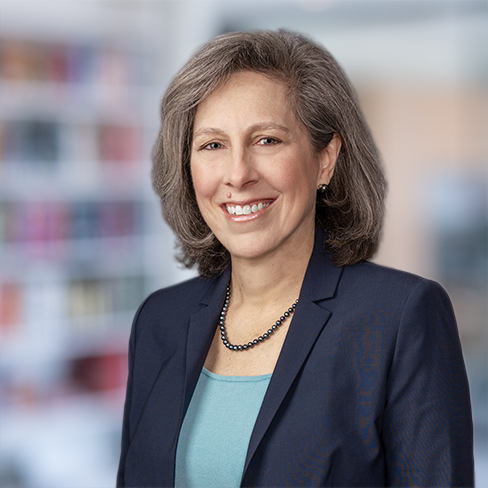 A woman with gray hair wearing a navy blazer, teal top, and pearl necklace smiles in an office setting with shelves in the background, reflecting the professionalism often seen in law offices or among lawyers in Chicago.