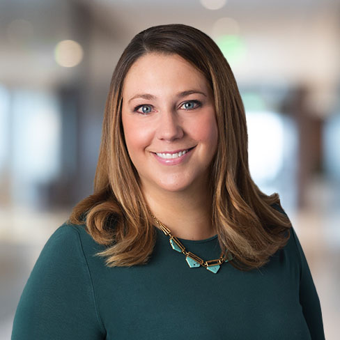 Woman with long brown hair wearing a dark green top and a statement necklace, smiling in a blurred corporate law office, showcasing the approachable side of lawyers in Chicago.