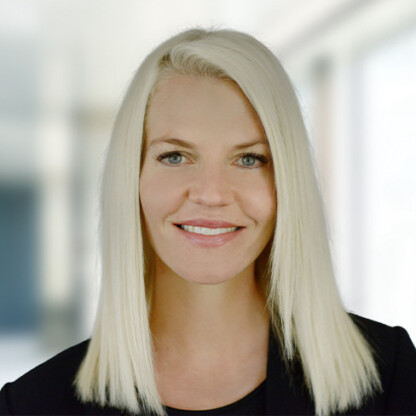 A woman with straight, shoulder-length blonde hair and light eyes, wearing a black top, is smiling at the camera in a brightly lit corporate law office.