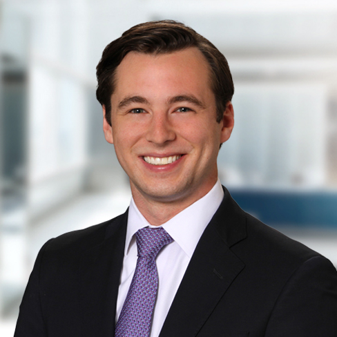 A man in a dark suit, white shirt, and purple tie smiles at the camera in a modern, bright corporate law office.