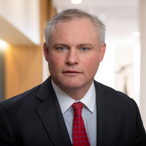 A middle-aged man in a dark suit and red tie poses for a professional headshot in a well-lit corporate law office hallway.