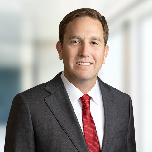 A man in a dark suit, white shirt, and red tie stands indoors against a blurred background in a corporate law office, looking at the camera and smiling.