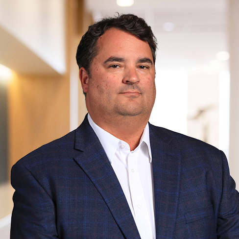 A man with dark hair wearing a blue checked suit jacket and white shirt stands in a brightly lit indoor setting, resembling the professional appearance often seen among Chicago lawyers in law offices.