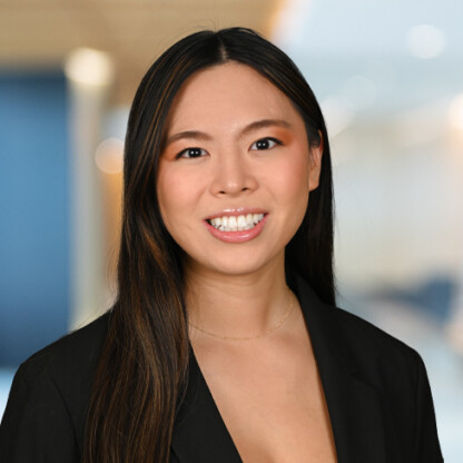 A woman with long straight hair, wearing a black blazer and a gold necklace, smiles at the camera in a brightly lit, blurred corporate law office setting.