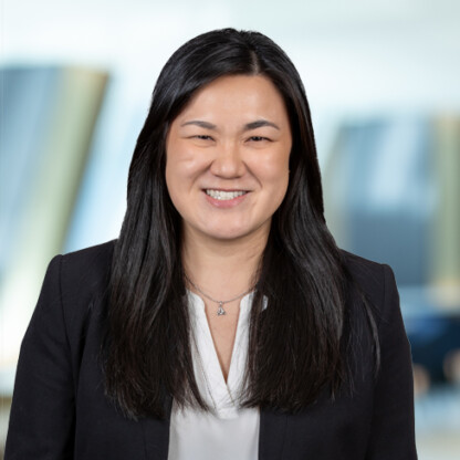 A woman with long dark hair, wearing a white blouse and dark blazer, smiles at the camera against a blurred office background typical of a corporate law office.