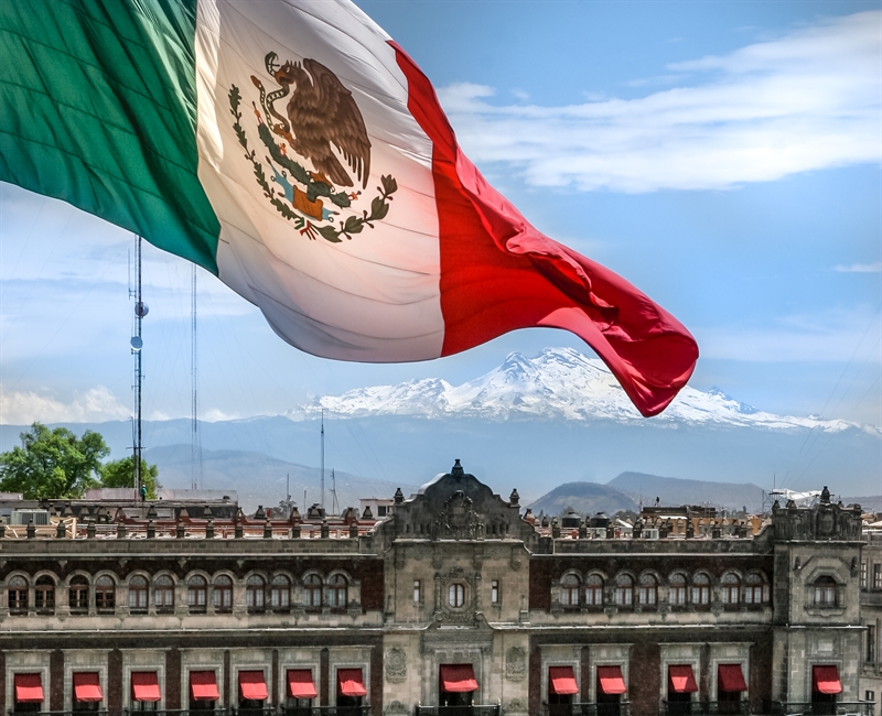 A large Mexican flag waves over historic buildings, with snow-capped mountains in the background and a prominent corporate law office under the clear blue sky.