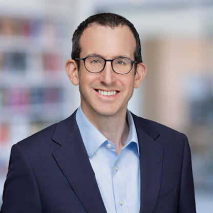 A man with short dark hair and glasses wearing a navy blazer and light blue shirt, smiling in a corporate law office setting.