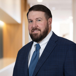A man with short dark hair and a beard, wearing a blue suit, white shirt, and blue tie, stands in a modern office hallway—reflecting the professionalism of lawyers in Chicago specializing in intellectual property law.