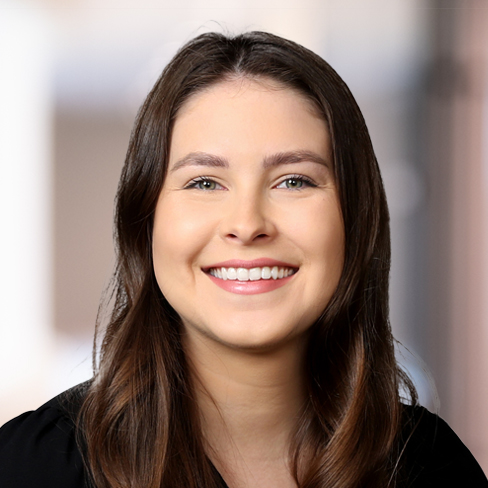A young woman with long brown hair smiles at the camera, wearing a black top, in front of a softly blurred corporate law office background.