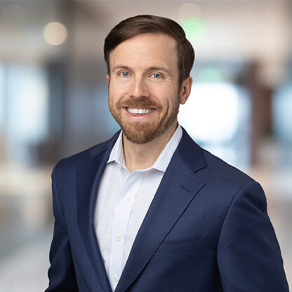 A man with brown hair and a beard, wearing a blue suit jacket and white shirt, smiles while standing in a blurred corporate law office setting.