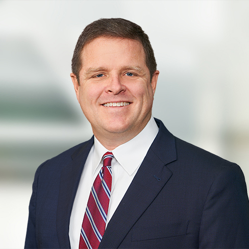 Man in a dark suit, white shirt, and red-striped tie smiling at the camera against a blurred light background, reflecting the professionalism of Chicago lawyers specializing in intellectual property law.