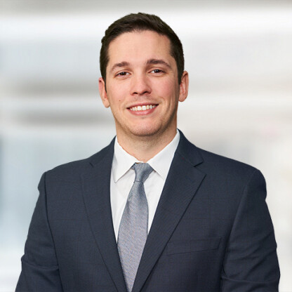 A man in a dark suit, white shirt, and light gray tie smiles at the camera against a blurred, light-colored background, reflecting the professionalism found at leading Chicago lawyers and corporate law office settings.