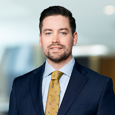 A man in a navy suit, light blue shirt, and yellow patterned tie stands in a corporate law office, looking at the camera with a neutral expression.