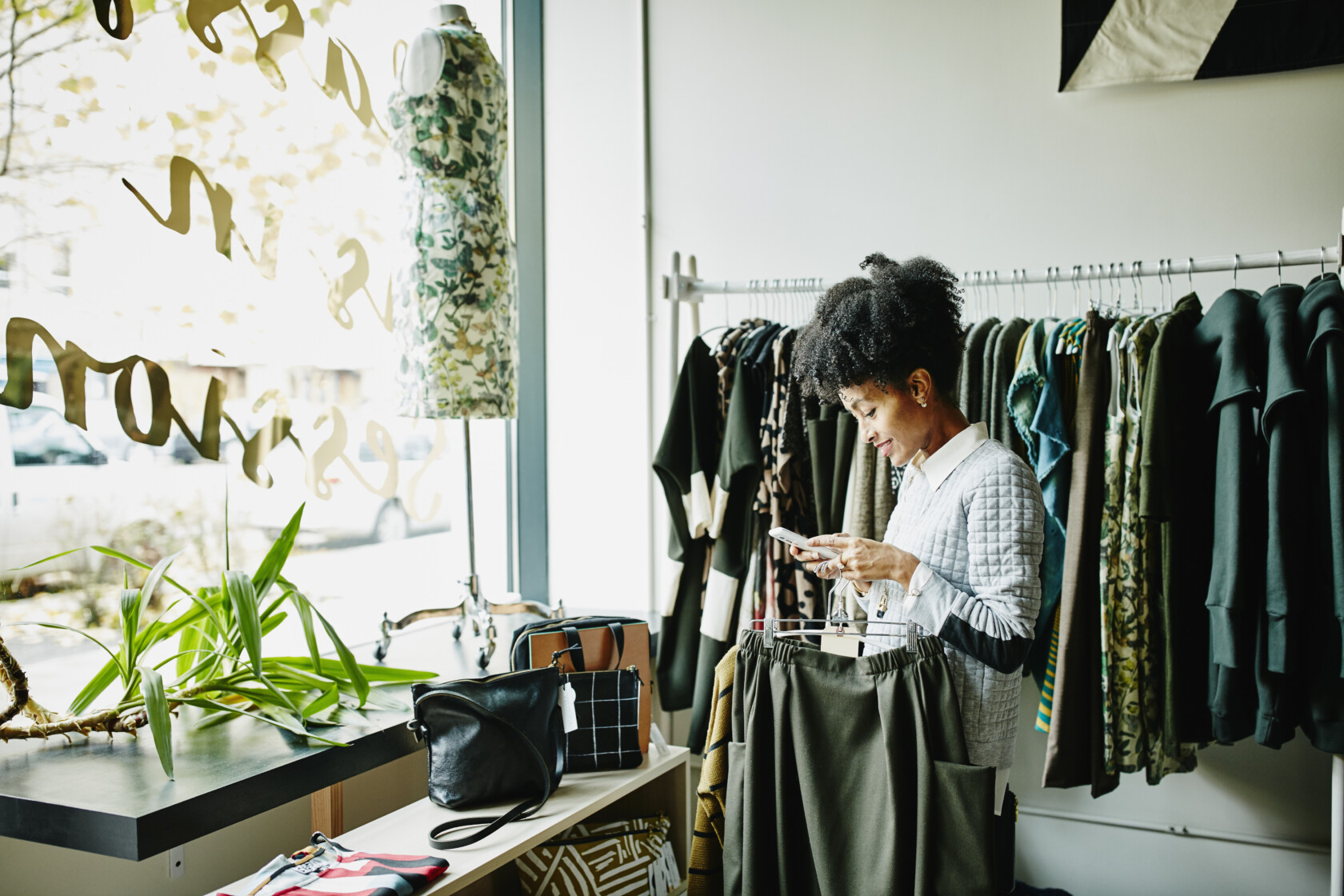 A person stands in a clothing store, looking at their phone while holding a pair of pants, perhaps searching for lawyers in Chicago, with racks of clothing and a mannequin visible in the background.