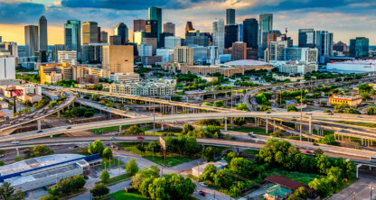 Aerial view of a city skyline with tall buildings, highways, and overpasses intersecting in the foreground—among them, prominent law offices providing litigation support—under a partly cloudy sky at sunset.