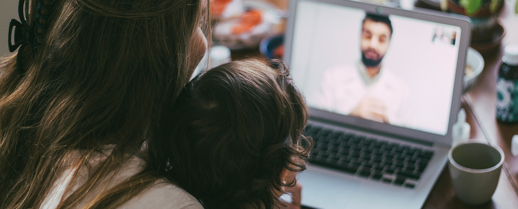 A woman and a child sit together facing a laptop, participating in a video call with a man from a corporate law office.
