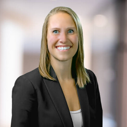 A woman with straight blonde hair, wearing a black blazer over a white top, smiles at the camera against a blurred corporate law office background.