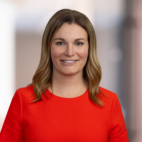 A woman with shoulder-length brown hair wearing a bright red top poses in front of a blurred indoor background, capturing the dynamic atmosphere of a Chicago lawyers' corporate law office.