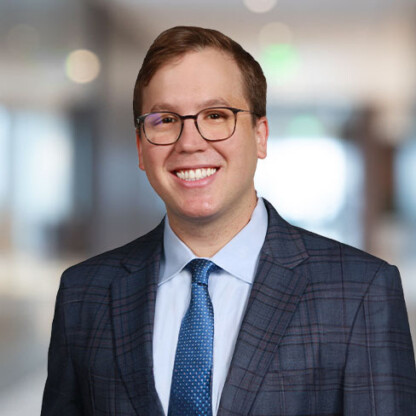 A man wearing glasses, a suit, and a blue tie smiles at the camera in a blurred corporate law office setting.