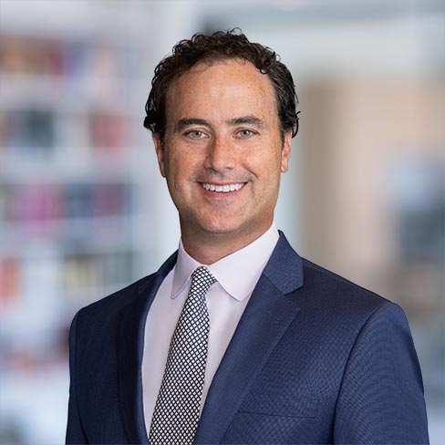 A man with curly dark hair wearing a navy suit, white shirt, and patterned tie stands smiling in front of a blurred office background at one of the leading law offices specializing in intellectual property law.