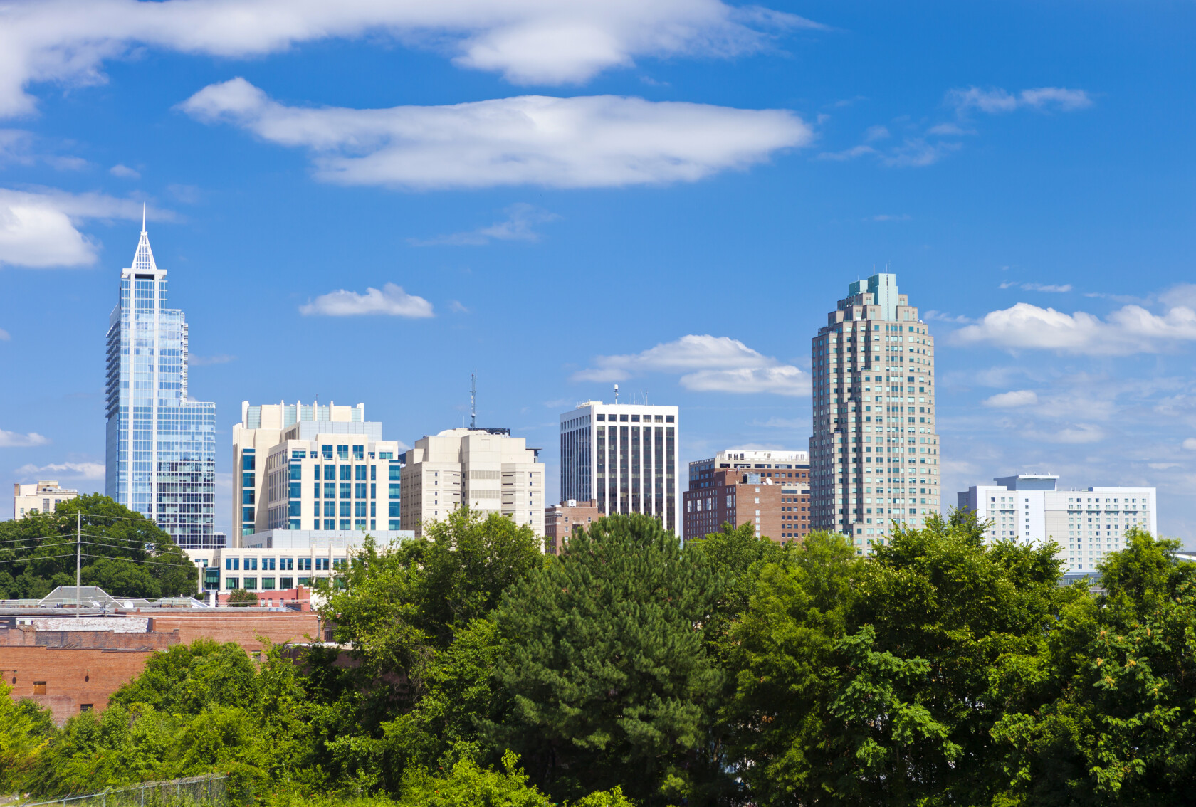 Raleigh city skyline with modern high-rise buildings, including a prominent corporate law office, and trees in the foreground under a blue sky with scattered clouds.