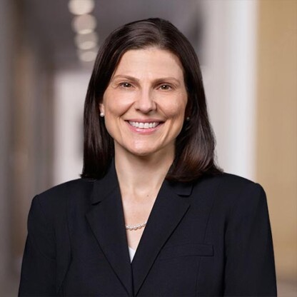A woman with straight dark hair, wearing a black blazer and pearl necklace, smiles while standing in a corporate law office hallway with blurred lights in the background.