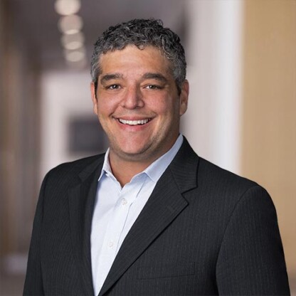 Smiling man with short curly hair wearing a dark suit jacket and light dress shirt, standing in a blurred indoor hallway of a corporate law office, representing Chicago lawyers.