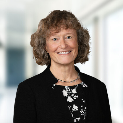 Smiling woman with curly brown hair wearing a black blazer and patterned blouse, standing in a brightly lit, blurred indoor setting, representing lawyers in Chicago.