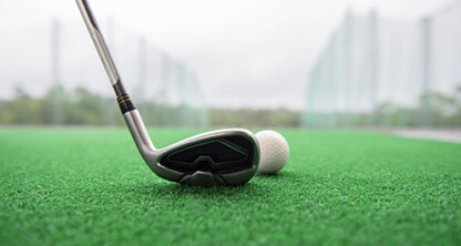 A close-up of a golf club positioned next to a golf ball on artificial green turf, with a blurred outdoor background—perfect imagery for law offices or corporate law office events in Chicago.