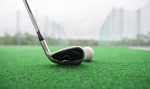 A close-up of a golf club positioned next to a golf ball on artificial green turf, with a blurred outdoor background—perfect imagery for law offices or corporate law office events in Chicago.