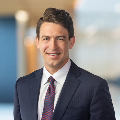 A man in a navy suit, white shirt, and red patterned tie stands indoors, smiling at the camera with a blurred law offices background, reflecting expertise in litigation support.