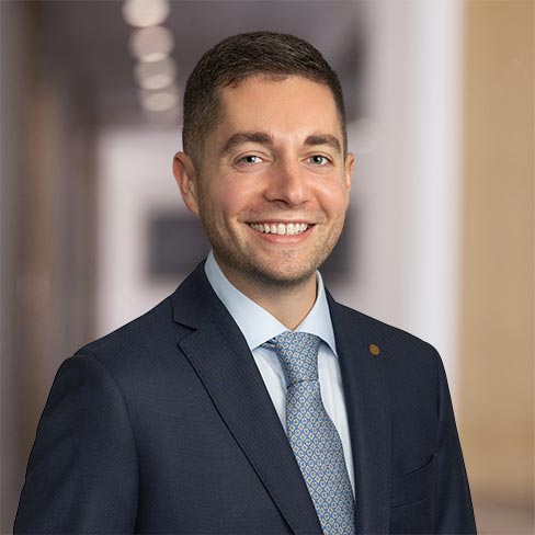 A man in a dark suit, light blue shirt, and patterned tie smiles at the camera while standing in a brightly lit hallway of a corporate law office.