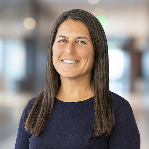 A woman with long straight brown hair, wearing a navy blue sweater, smiles at the camera in a brightly lit, blurred corporate law office.