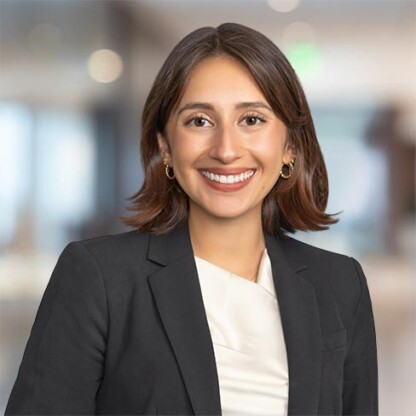 A woman with short brown hair, wearing a black blazer and white top, smiles at the camera in a modern corporate law office with a blurred background.